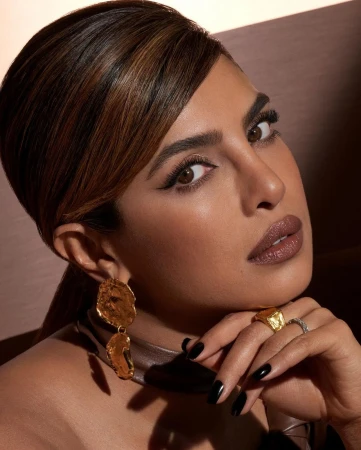 Close-up portrait of Priyanka Chopra wearing textured gold earrings, a gold ring, and dark nail polish with her hand beneath her chin