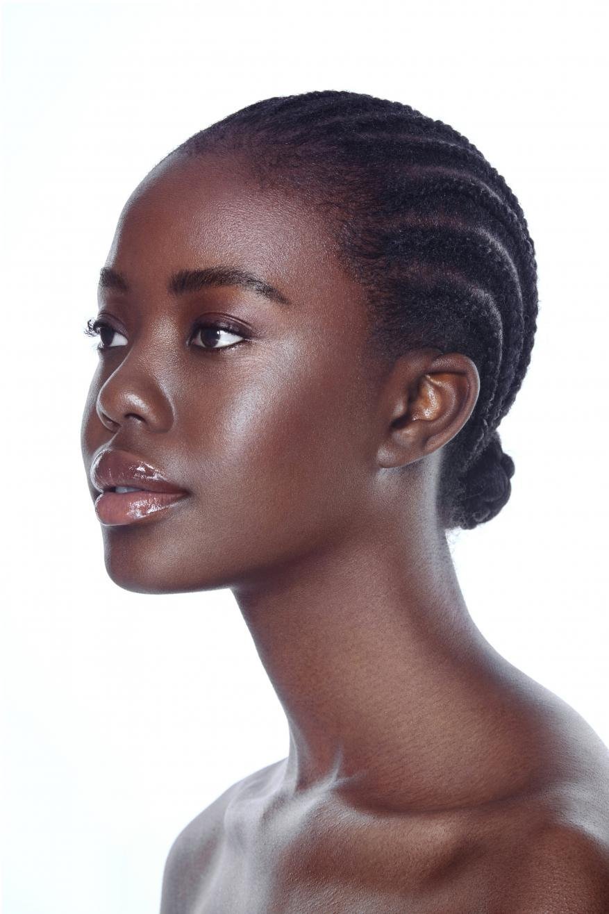 Headshot of a dark-skinned woman with braided hair in profile against a white background. Beauty shot with glossy makeup.
