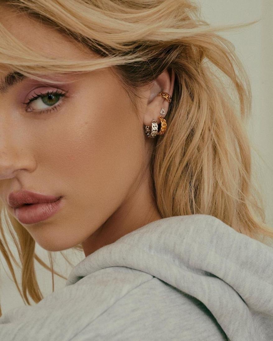 Close-up portrait of a blonde woman with hoop earrings and a grey hoodie, focusing on her face, hairstyle, and fashion accessories.