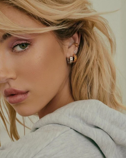 Close-up portrait of a blonde woman with hoop earrings and a grey hoodie, focusing on her face, hairstyle, and fashion accessories.