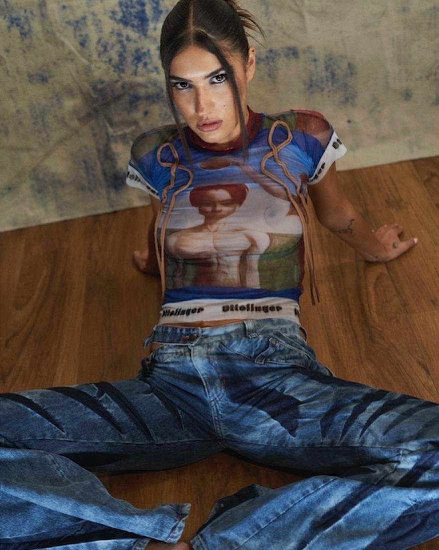 Woman in a printed T-shirt and jeans sits on the floor with an intense gaze; fashion, casual style, indoor setting.