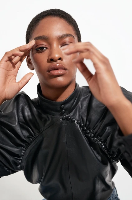 Close-up shot of a young adult woman with short hair wearing a black leather-like blouse with decorative buttons, posing in a photo studio.