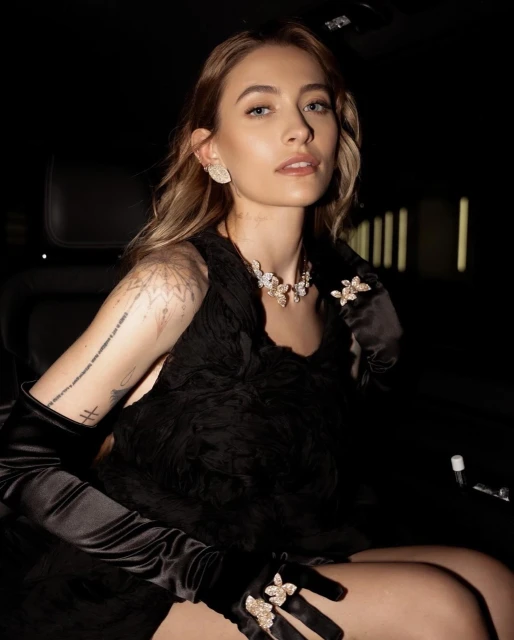 Woman in a black dress with diamond jewelry posing indoors in front of a black car seat and window. The shot is eye-level.