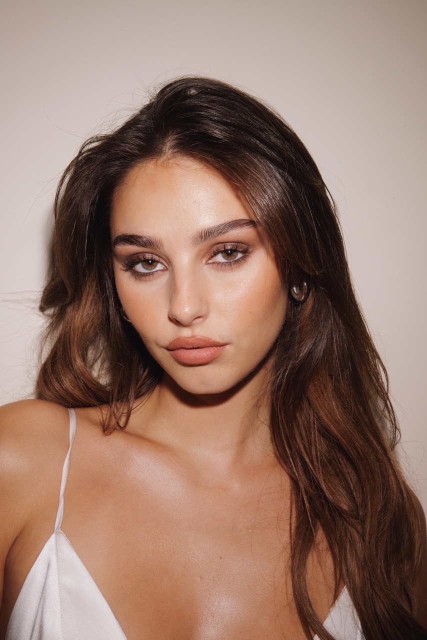 Close-up portrait of a young woman with long brown hair, wearing a white top, posing in front of a neutral-toned background.