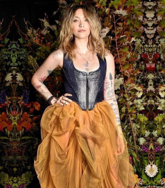 Full shot of a young adult woman in a sleeveless top with a gold tulle skirt posing against a backdrop of autumnal foliage.
