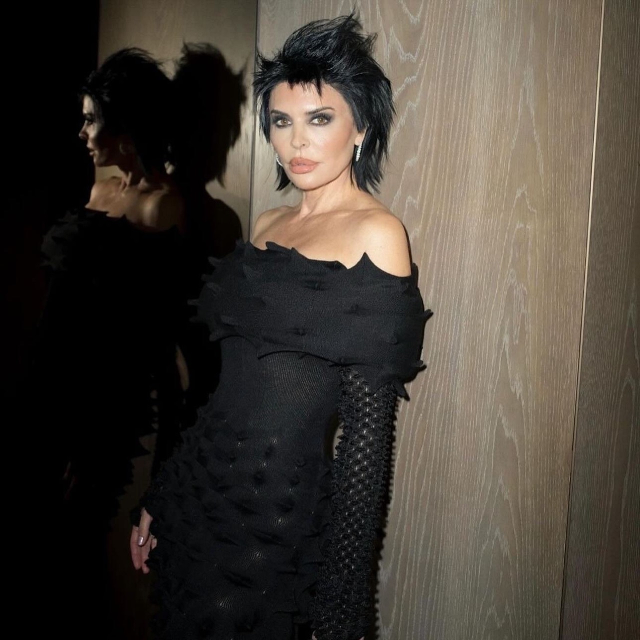 Close-up of Lisa Rinna in a textured, off-the-shoulder black dress, with a dark hairstyle and a reflective background, posing indoors.