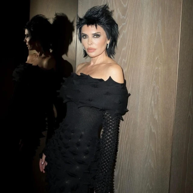 Close-up of Lisa Rinna in a textured, off-the-shoulder black dress, with a dark hairstyle and a reflective background, posing indoors.