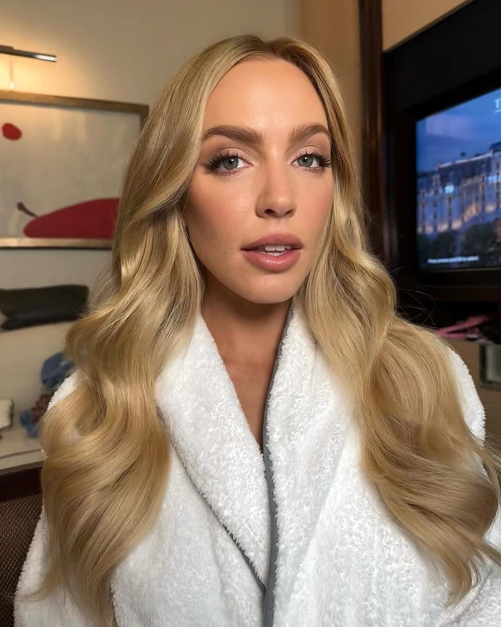 Close-up portrait of a woman with blonde hair wearing a white plush robe against an indoor hotel room background.