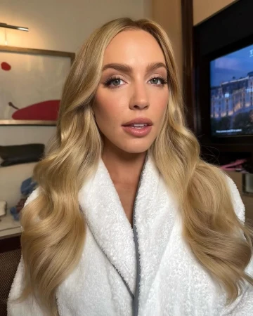 Close-up portrait of a woman with blonde hair wearing a white plush robe against an indoor hotel room background.