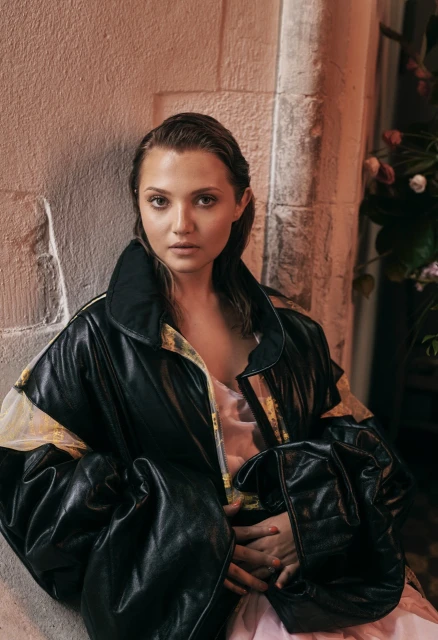 Portrait of a young woman with wet hair wearing a black jacket, posed against a textured wall with a flower arrangement in the background.