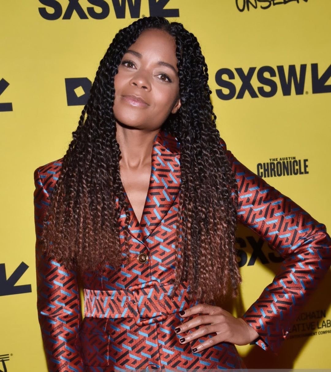 Portrait of an adult female with dark hair in patterned blazer at a SXSW event, posing with hand on hip against a yellow backdrop.