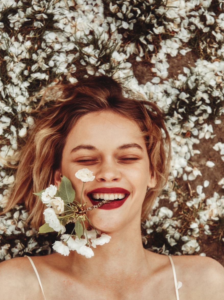 Smiling woman lying amidst scattered white flower petals holding a small flowering twig in her mouth