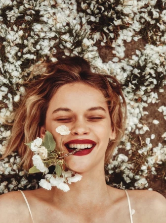 Smiling woman lying amidst scattered white flower petals holding a small flowering twig in her mouth