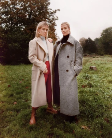 Two women posing outdoors on grass in an open field wearing long winter coats, with makeup by Jacinta Spencer.