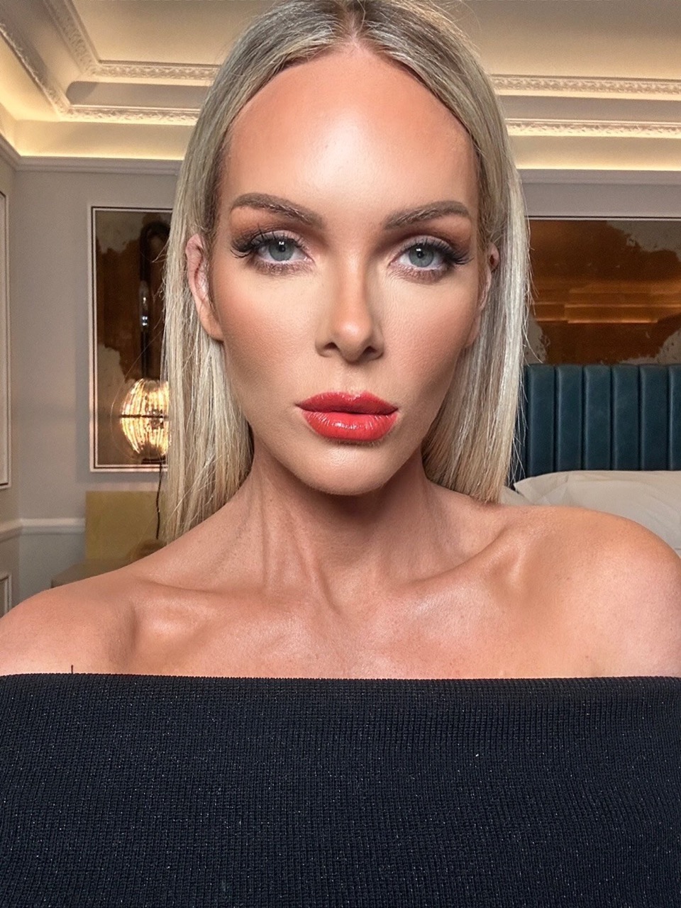 Close-up of an adult female posing indoors with blonde hair, orange lipstick, dark eye makeup, and a black off-the-shoulder top.
