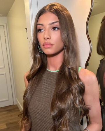 Portrait of a woman with long brown hair wearing a textured olive top and earrings, posed indoors near a mirror.