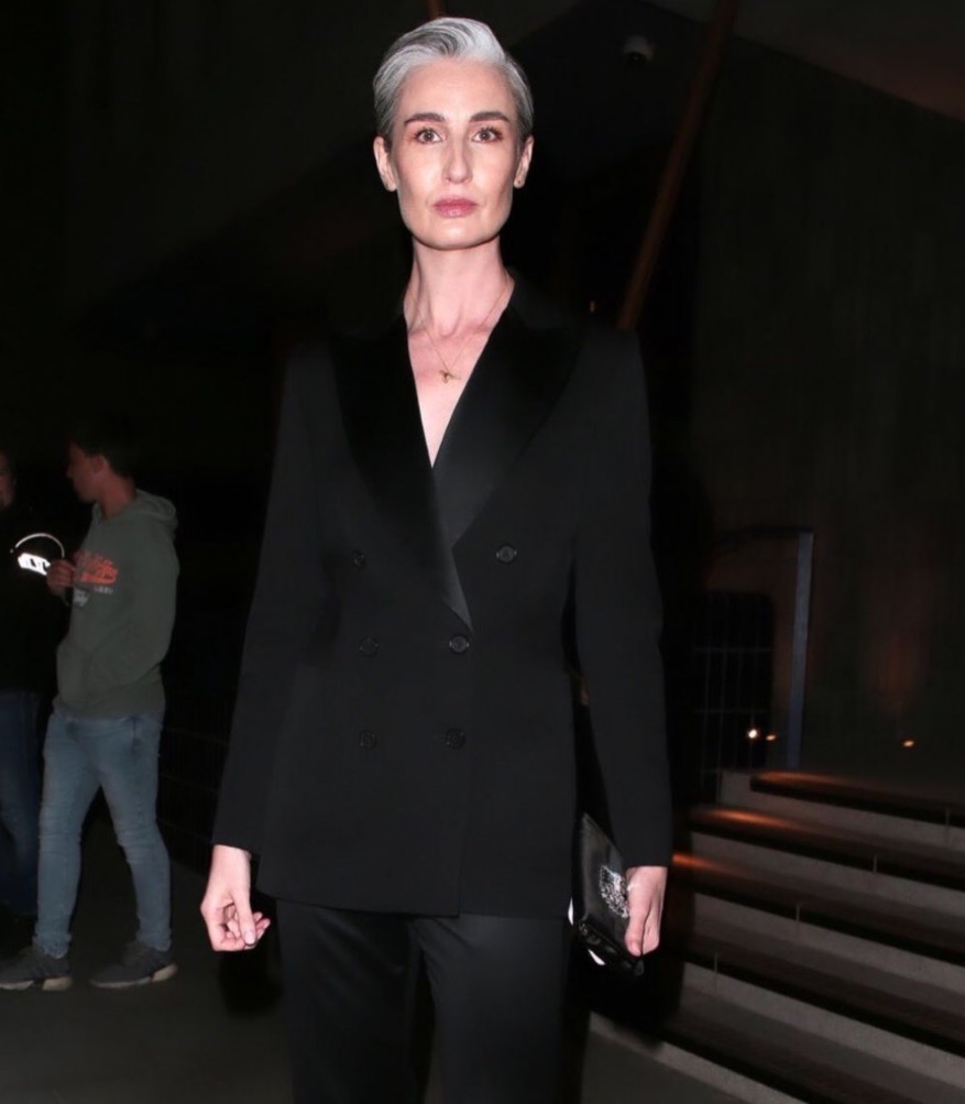 Elegant adult woman in a black tuxedo suit and short grey hair posing indoors, with two people visible in the background.
