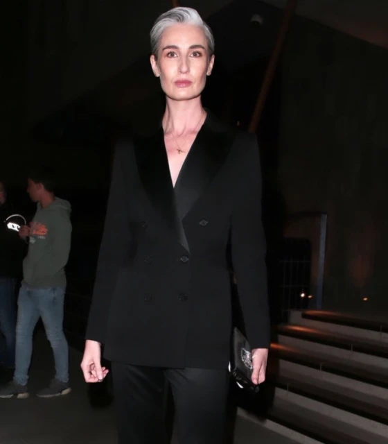 Elegant adult woman in a black tuxedo suit and short grey hair posing indoors, with two people visible in the background.
