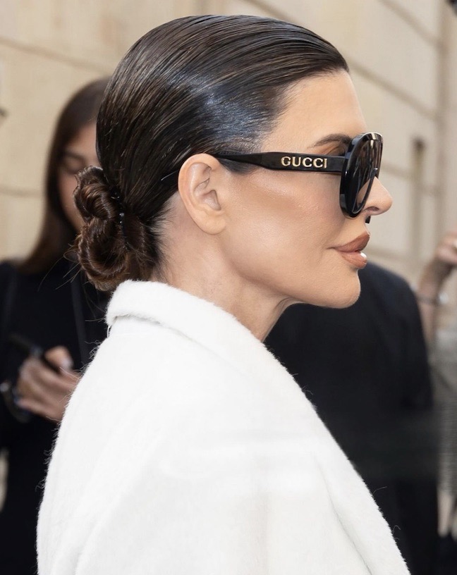 Adult woman in profile wearing Gucci sunglasses with a low bun hairstyle and a white coat, standing outdoors in soft lighting.