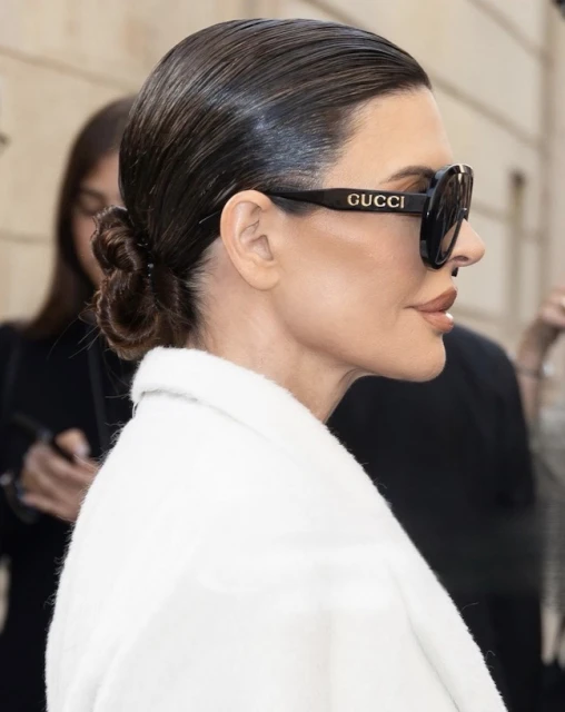 Adult woman in profile wearing Gucci sunglasses with a low bun hairstyle and a white coat, standing outdoors in soft lighting.