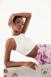 Black woman wearing a white crop top and pink tie-dye skirt posing on a metallic flight case against a plain white background