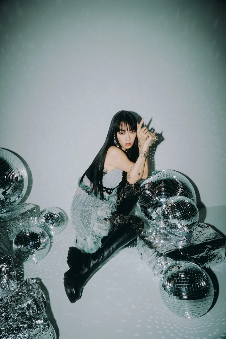 Portrait of a woman with long black hair and tattoos, posed among silver disco balls in a photo studio with white seamless backdrop.
