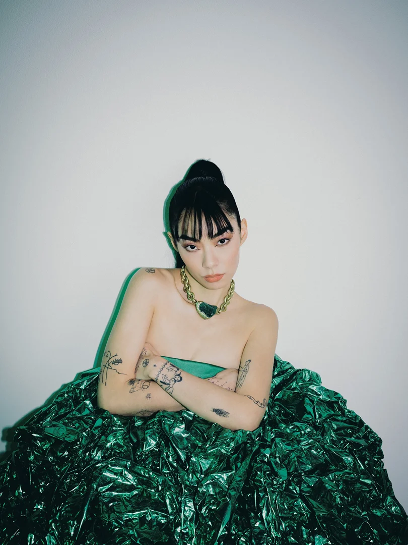 Portrait of a young adult woman with a high bun and arm tattoos wearing a green dress and necklace against a white background.