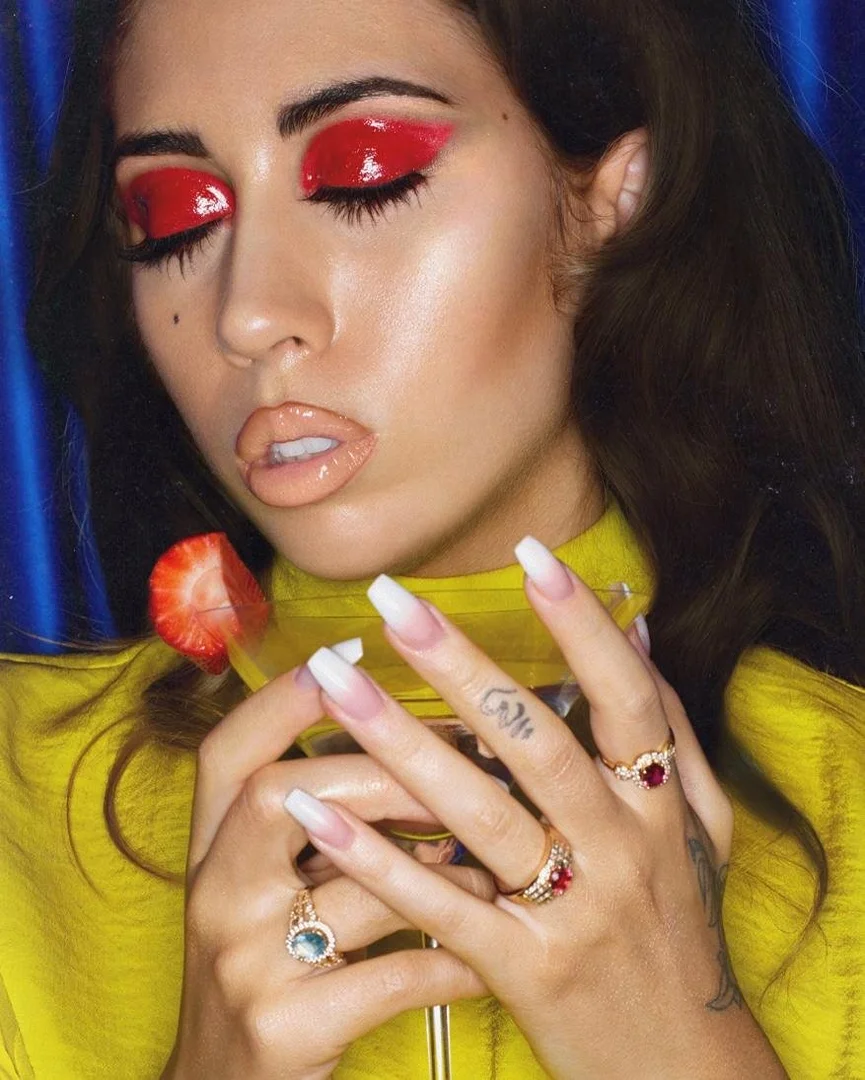 Close-up of a stylish adult female with red eye makeup holding a cocktail glass with a strawberry, showcasing fashion and beauty.