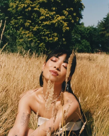 Woman with dark hair and tattoos sitting in a tall golden grass field with trees in the background