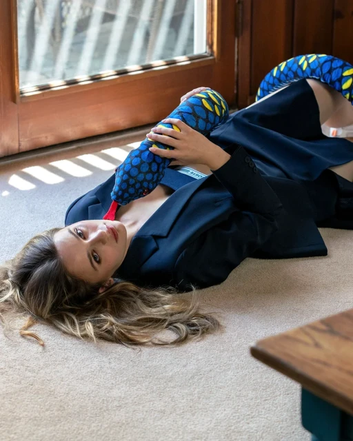 Attractive woman in blazer lies on floor with blue snake plush toy near open glass doors with natural light and neutral tones.