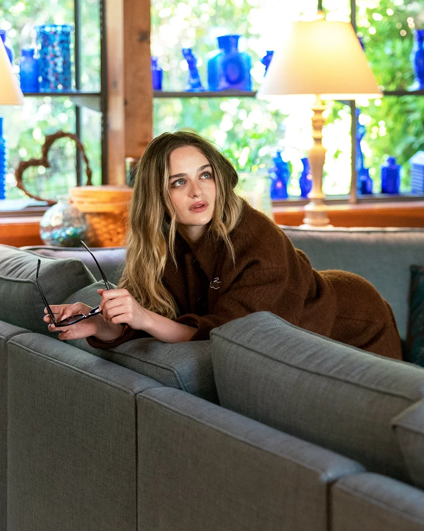 Attractive adult woman with long wavy hair poses on a grey couch with blue glass vases in the background and wearing a brown sweater.