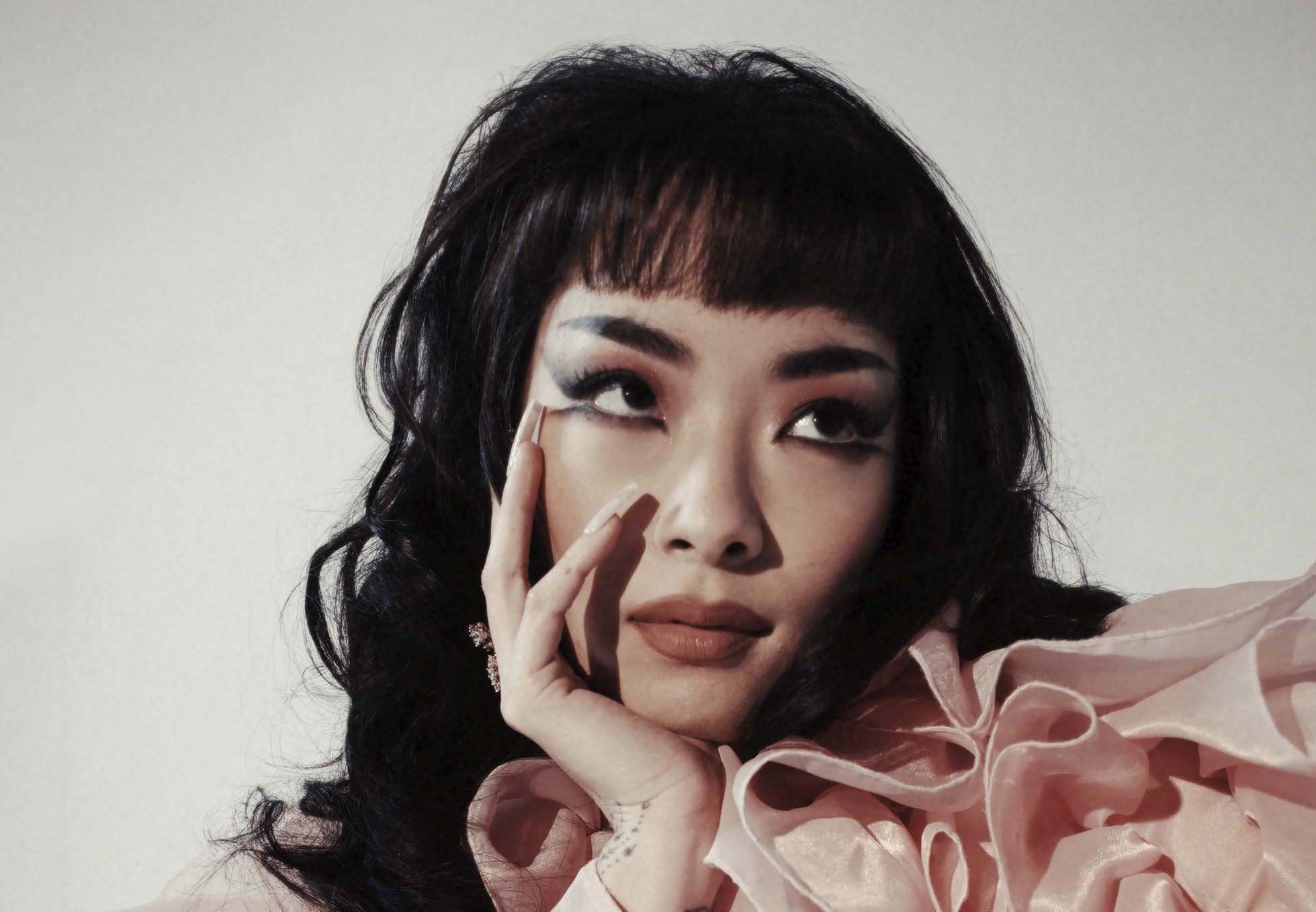 Close-up portrait of a woman with dark hair, bangs, dramatic graphic eyeliner, and a pink ruffled collar.