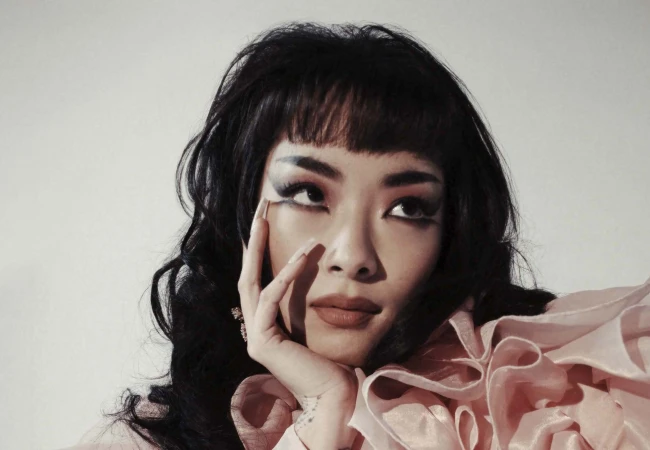 Close-up portrait of a woman with dark hair, bangs, dramatic graphic eyeliner, and a pink ruffled collar.