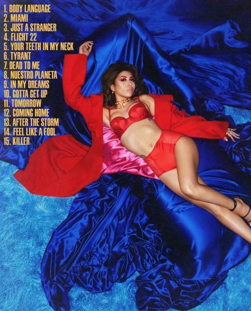 Woman in red lingerie and coat poses on blue silk fabric, lying down and looking at the camera. List of song titles on left side.