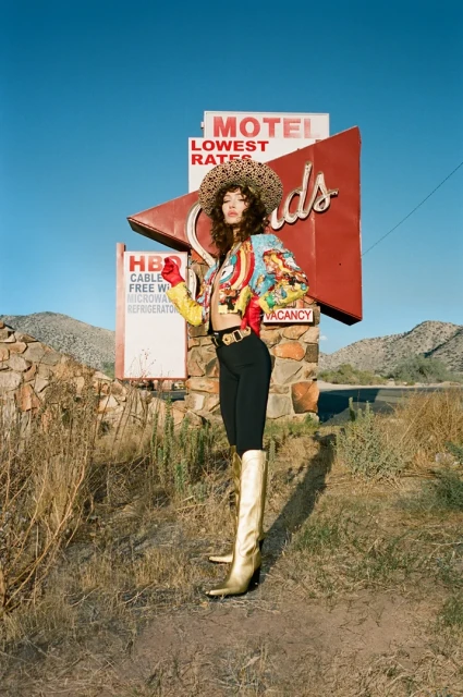 A woman stands in a desert landscape wearing a colorful jacket, black pants, gold boots, and a leopard print hat with a vintage motel sign.