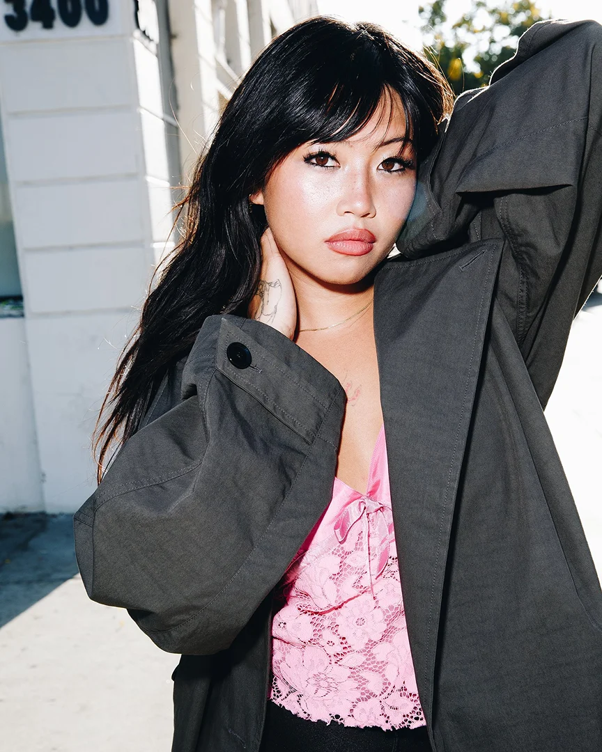 Portrait of a young woman with long dark hair posing outdoors wearing a dark green coat and pink floral lace top, looking at camera.