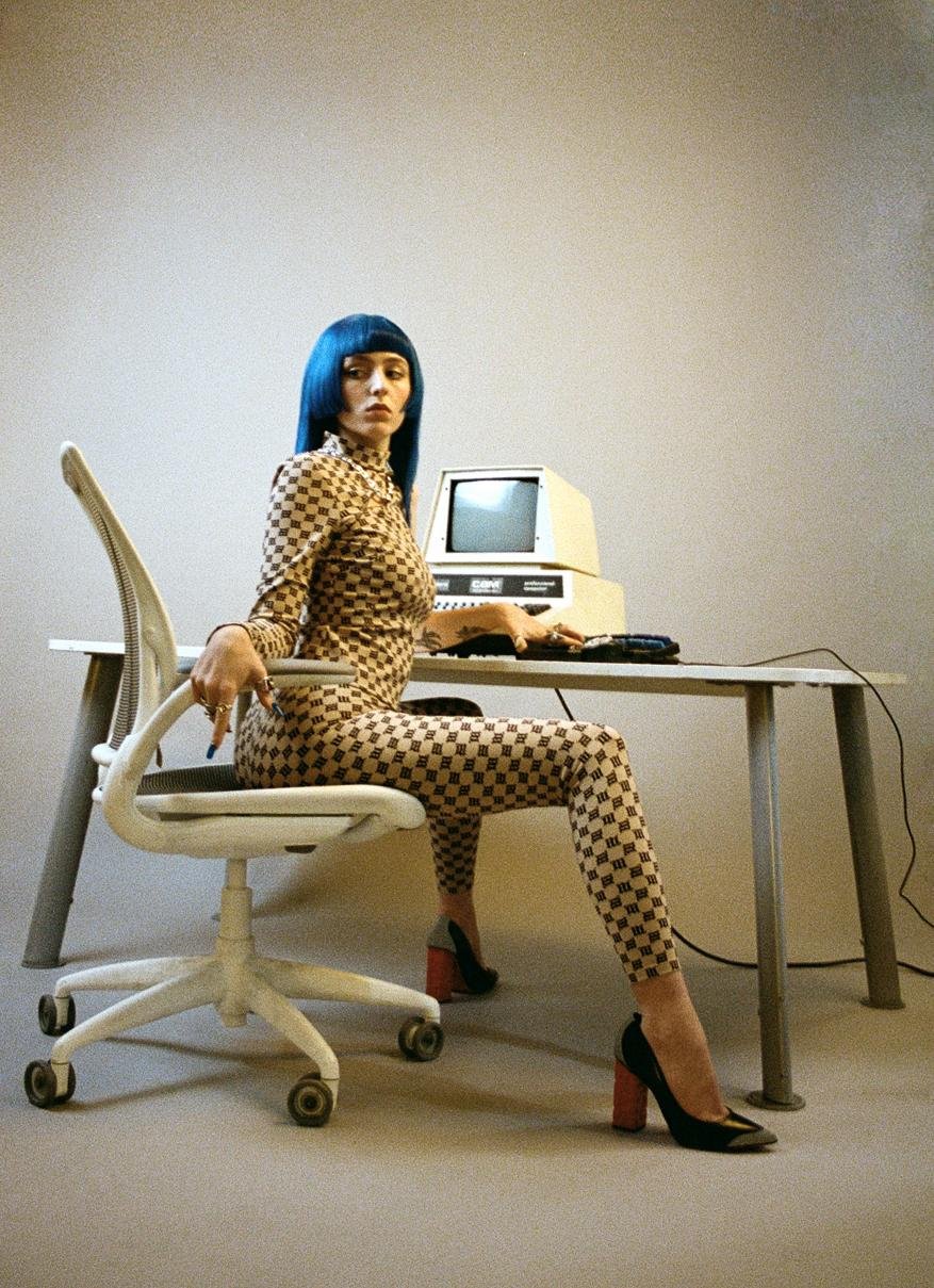 Woman with blue hair sits at a desk with an old computer, wearing a checkered jumpsuit and high heels in a bright, neutral studio.