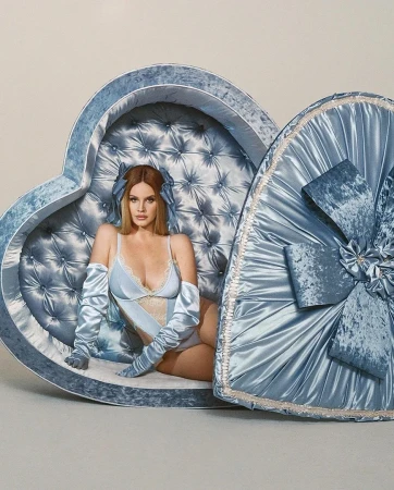 Lana Del Rey poses in a blue lingerie set and long gloves inside of a plush, heart-shaped chair against a neutral background.
