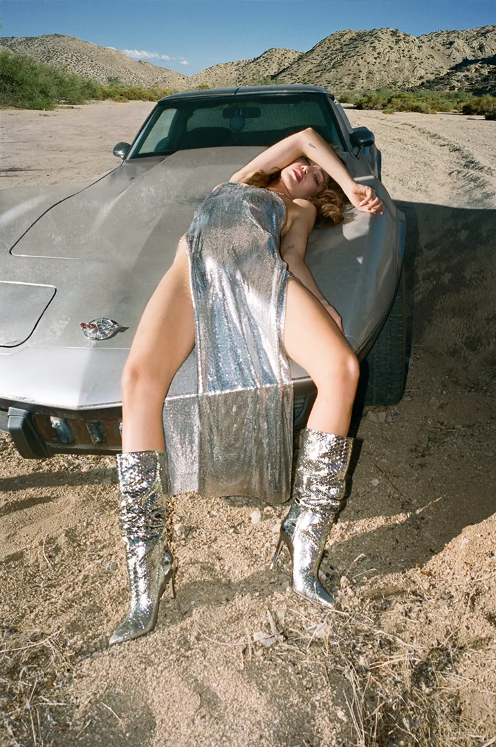 Woman in a silver dress and boots poses on a vintage car in the desert with mountains in the background on a sunny day.