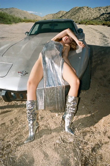 Woman in a silver dress and boots poses on a vintage car in the desert with mountains in the background on a sunny day.