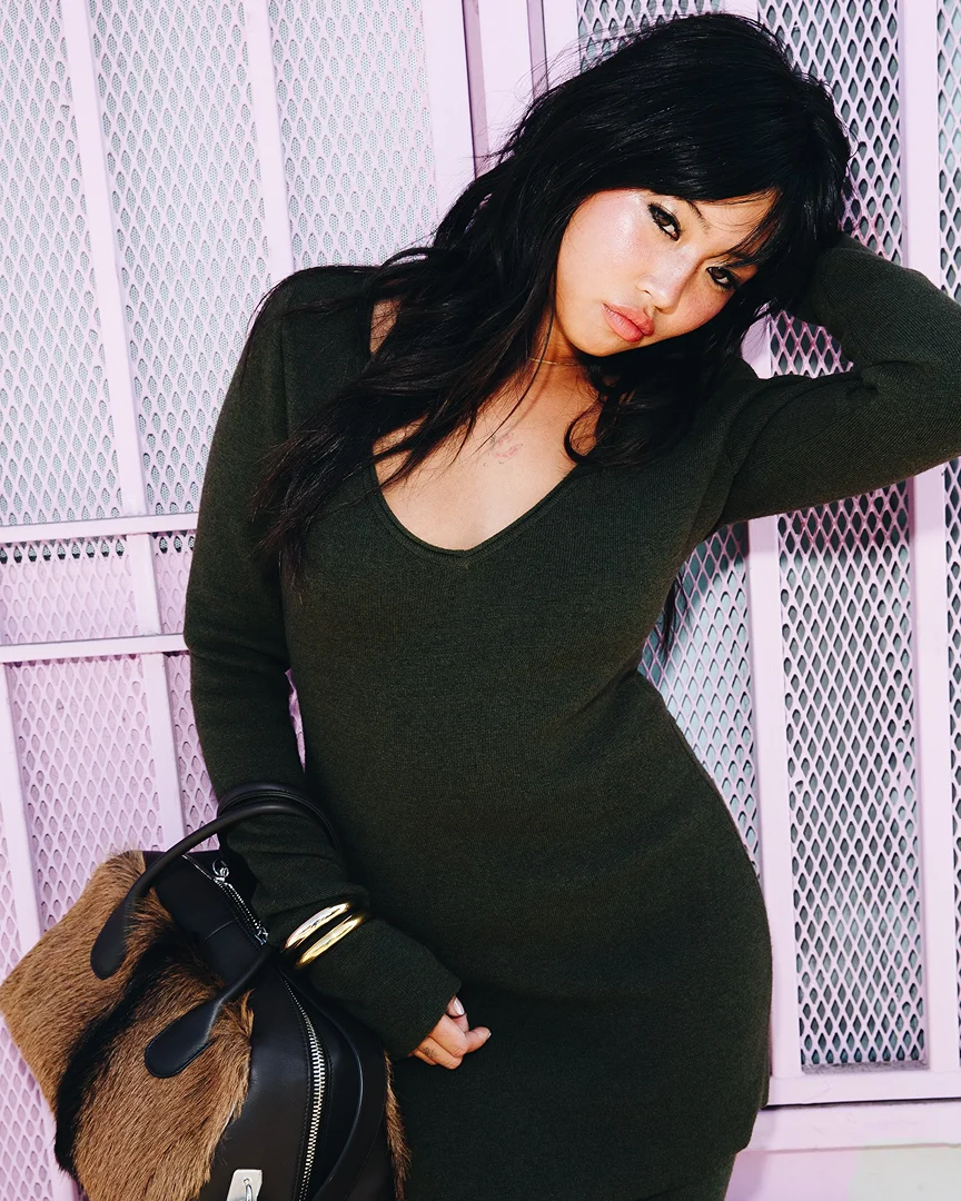 A fashionable woman in a green sweater dress poses against a pink diamond-patterned wall, holding a brown and black fur handbag.
