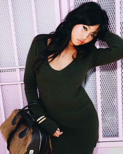 A fashionable woman in a green sweater dress poses against a pink diamond-patterned wall, holding a brown and black fur handbag.