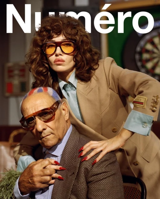 Fashion magazine cover featuring a woman in a tan suit standing behind an older man in glasses, with a dartboard in the background.