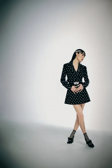 A stylish adult woman with long hair in a black polka dot blazer dress and matching shoes is posing in a bright photo studio.