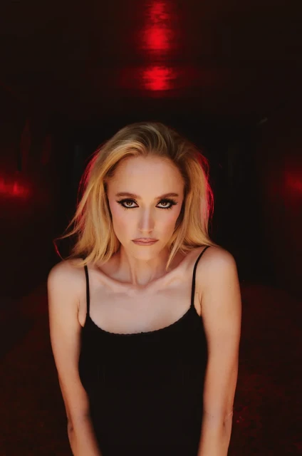 Blonde woman in a black tank top gazes intensely at the camera, dramatic makeup, against a dark background with moody red lighting.