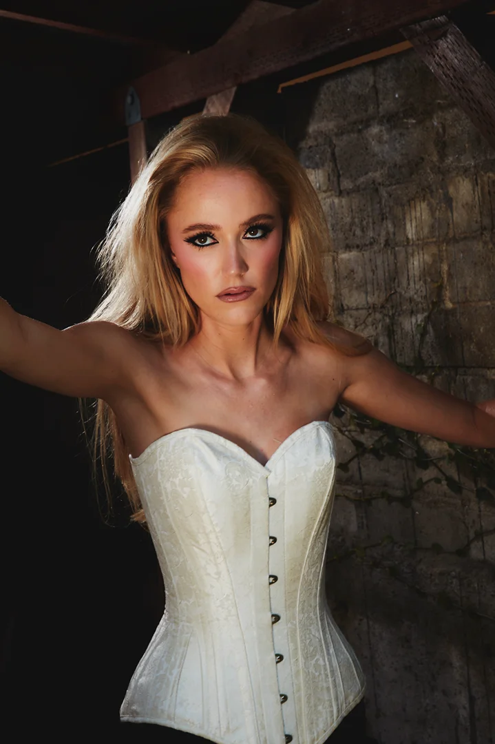Blonde woman in corset poses indoors against a stone wall with wooden beams. Low key lighting for fashion or beauty shot.