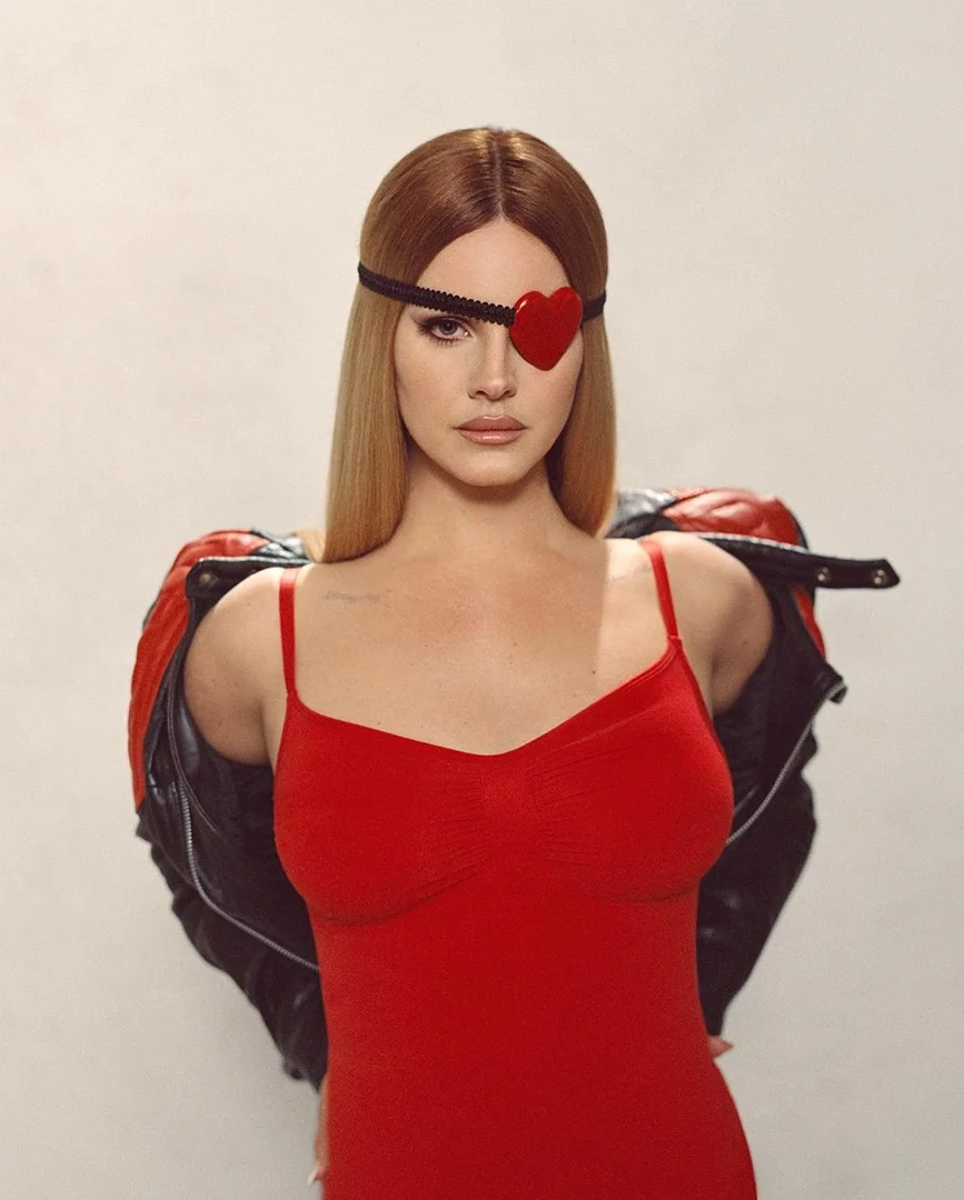 Portrait of a blonde woman wearing a red dress and heart-shaped eyepatch, with a black leather jacket draped over her shoulders.