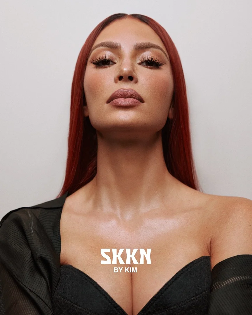 Glamour portrait of Kim Kardashian with red hair wearing a black top against a white backdrop, showcasing beauty and fashion elements.