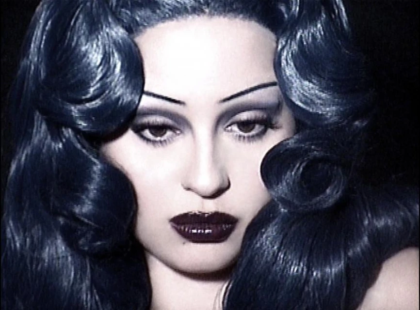 Glamorous close-up portrait of a woman with dramatic makeup, including dark lipstick, arched eyebrows, and voluminous dark curly hair.