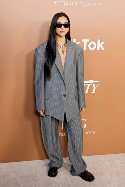 Dua Lipa in a grey pinstripe suit and sunglasses poses against a tan backdrop featuring brand logos at a promotional event.